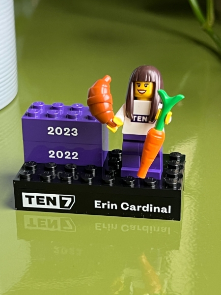 A Lego mini-figurine with long brown hair, holding a croissant and a carrot. It is standing on a Lego platform that says TEN7 and Erin Cardinal plus two bricks for the years 2022 and 2023.