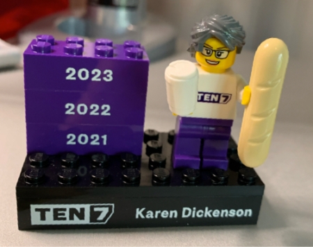 A Lego mini-figurine with grey hair and glasses, holding a croissant and a coffee mug, on a platform that says TEN7 and Karen Dickenson plus three bricks for the years 2021, 2022 and 2023