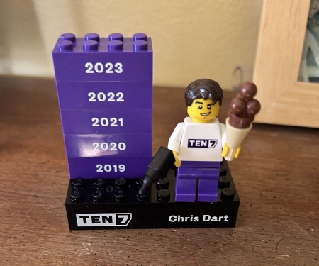 A Lego mini-figurine with short brown hair, holding a drill and a chocolate ice cream cone, on a platform that says TEN7 and Chris Dart plus 5 bricks for the years 2019-2023