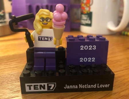 A Lego mini-figurine with blonde hair and glasses, holding a paint roller and a strawberry ice cream cone, on a platform that says TEN7 and Janna Netland Lover plus two bricks for the years 2022 and 2023