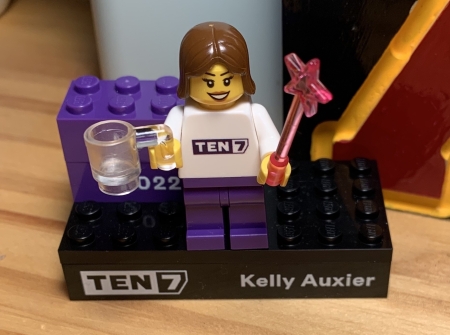 A Lego mini-figurine with brown hair, holding a clear mug and a magic wand, on a platform that says TEN7 and Kelly Auxier plus two bricks for the years 2022 and 2023