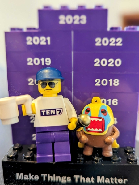 A Lego mini-figurine with a blue hat and sunglasses on, holding a white mug and a microphone, next to a Domo in a mask, on a platform that says Make Things That Matter plus 13 bricks for the years 2011-2023