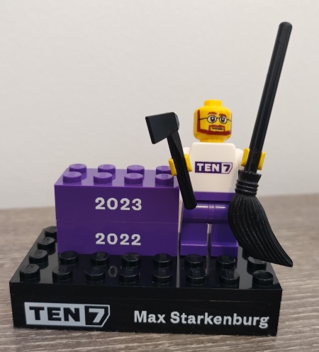 A Lego mini-figurine with glasses and a beard, holding an axe and a broom, on a platform that says TEN7 and Max Starkenburg plus two bricks for the years 2022 and 2023