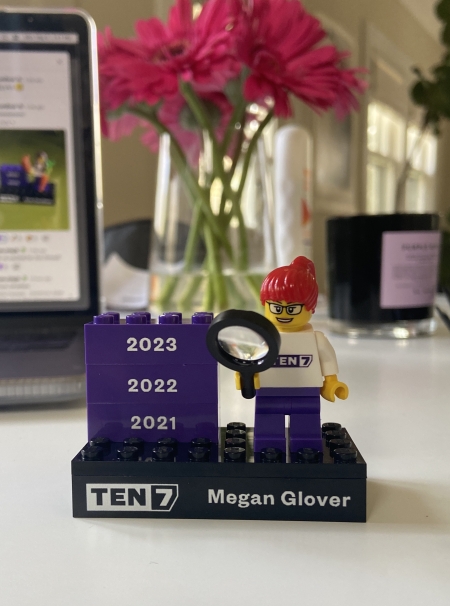 A Lego mini-figurine with a red ponytail, holding a magnifying glass, on a platform that says TEN7 and Megan Glover plus three bricks for the years 2021, 2022 and 2023